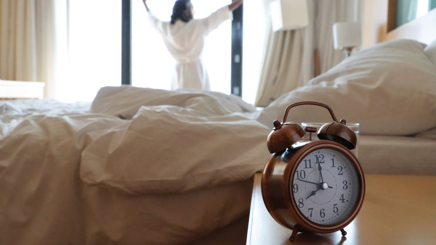 Stock photo showing Indian man wearing white towelling robe stood stretching in front of hotel window with open curtains looking out of window with a close-up view of retro, double bell, analogue alarm clock sitting on bedside table.